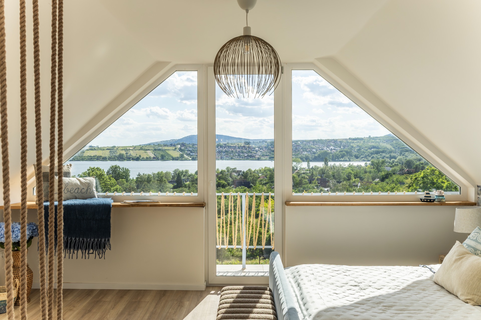 Bright attic bedroom with triangular windows, lake view, hanging woven pendant lamp, and cozy bedding.